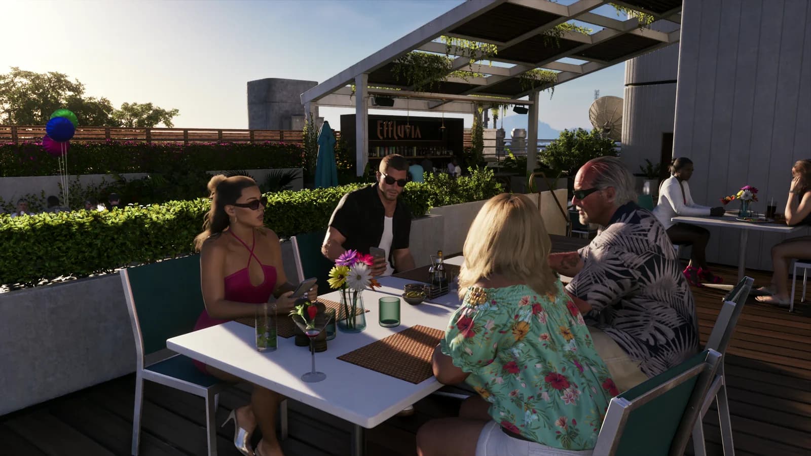 Il gruppo di quattro persone è seduto a un tavolo con fiori e bicchieri da cocktail; dietro di loro, sopra il bancone del bar, si legge chiaramente l'insegna "Effluvia".