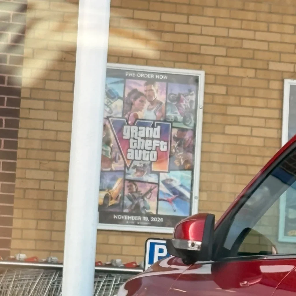 Il poster di GTA VI sulla facciata del negozio, visto dal parcheggio con un’auto in primo piano.